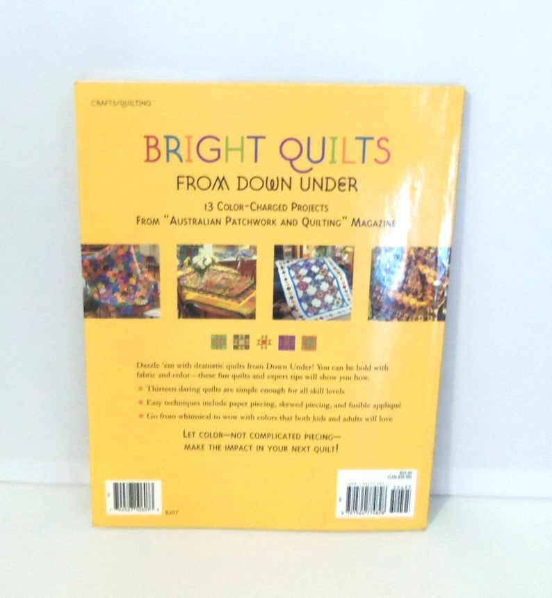 May include: A yellow softcover book titled Bright Quilts from Down Under, featuring 13 color-charged projects from Australian Patchwork and Quilting magazine. The back cover displays four small quilt photos, text descriptions, and a barcode.