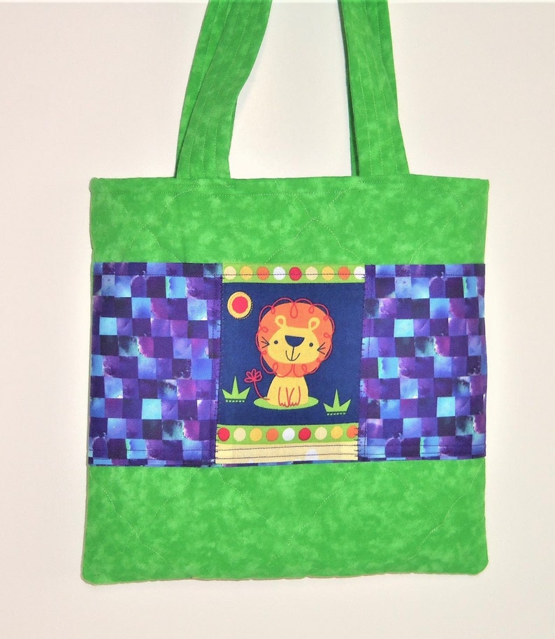 May include: A quilted green tote bag featuring two long handles and a central horizontal panel. The panel displays a dark blue section with a cartoon lion illustration, flanked by two sections of a purple and blue checkered pattern fabric.