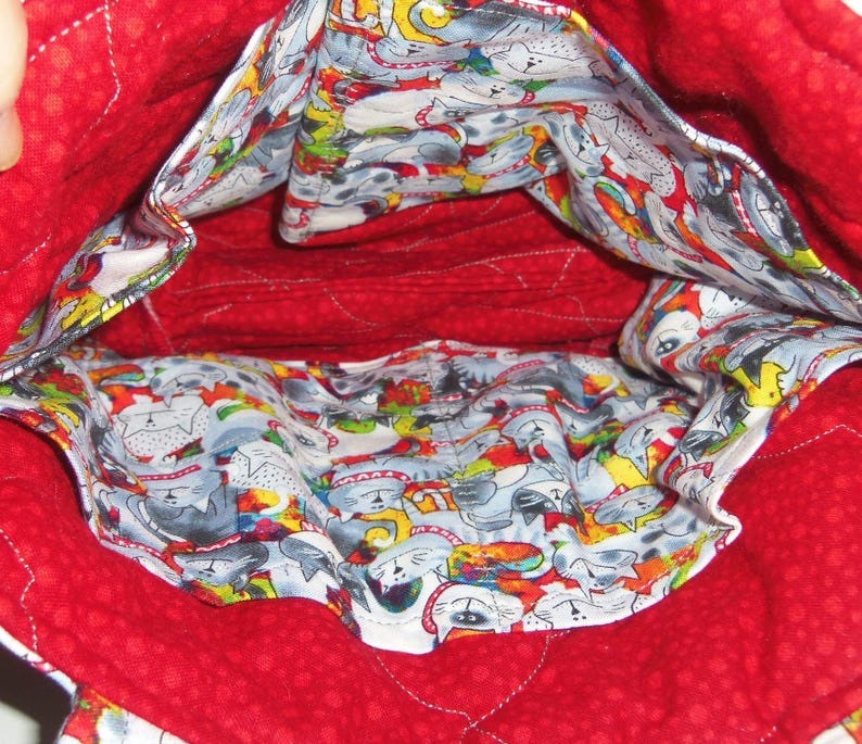 May include: A quilted fabric bag interior featuring a bright red lining with a subtle tonal dot pattern. The interior pockets are lined with a white fabric printed with a colorful, repeating pattern of stylized cats in various poses.
