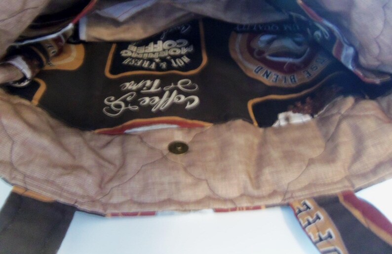 Puede incluir: Un bolso de tela con forro interior marr&oacute;n que presenta un patr&oacute;n acolchado y un cierre central de broche met&aacute;lico. La tela interior muestra un estampado con tem&aacute;tica de caf&eacute; con textos como Coffee Time, Hot & Fresh Morning Coffee y House Blend.