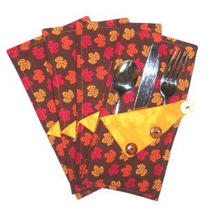 May include: Set of four brown fabric napkins with a red and orange leaf pattern. The napkins have a yellow pocket on the side to hold a fork, knife, and spoon.