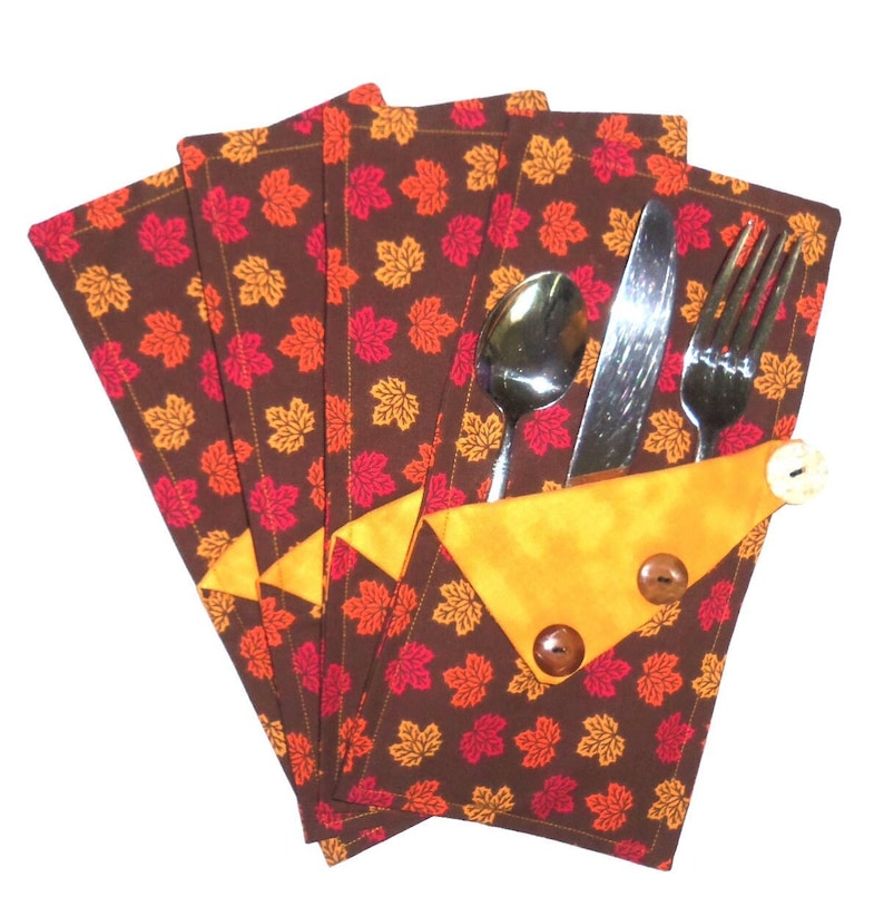 May include: Set of four brown fabric napkins with a red and orange leaf pattern. The napkins have a yellow pocket on the side to hold a fork, knife, and spoon.