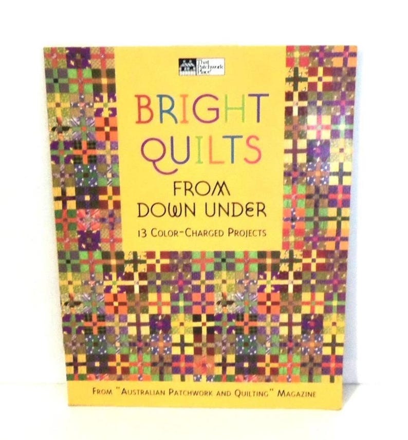 May include: A softcover book titled Bright Quilts From Down Under: 13 Color-Charged Projects From Australian Patchwork and Quilting Magazine. The cover features a yellow center with multicolored patchwork quilt patterns on the sides and bottom.