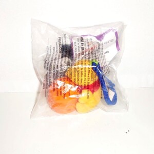 K&ouml;nnte beinhalten: Ein versiegelter, durchsichtiger Plastikbeutel mit einem kleinen, bunten Spielzeug, das aus einer leuchtend orangefarbenen, kugelf&ouml;rmigen Basis und einer gelben, abgerundeten Figur besteht. Ein blauer Kunststoffclip ist seitlich angebracht. Die Verpackung enth&auml;lt gedruckten Text auf Englisch, Franz&ouml;sisch und Spanisch.