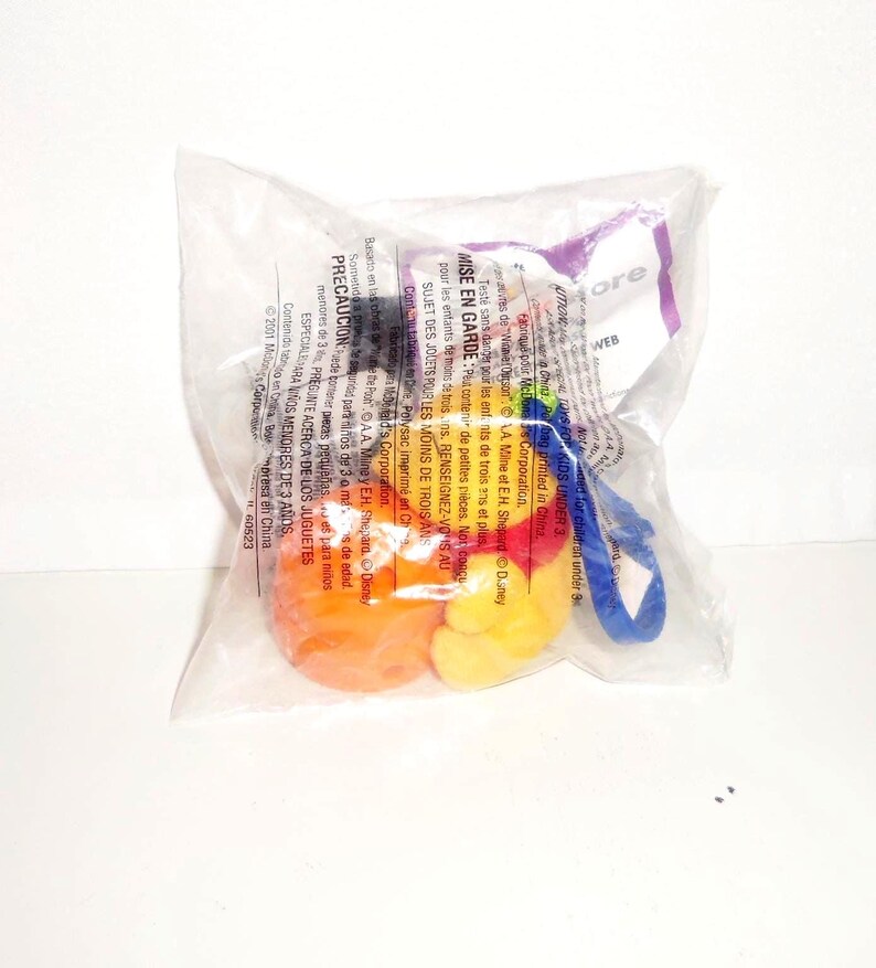 K&ouml;nnte beinhalten: Ein versiegelter, durchsichtiger Plastikbeutel mit einem kleinen, bunten Spielzeug, das aus einer leuchtend orangefarbenen, kugelf&ouml;rmigen Basis und einer gelben, abgerundeten Figur besteht. Ein blauer Kunststoffclip ist seitlich angebracht. Die Verpackung enth&auml;lt gedruckten Text auf Englisch, Franz&ouml;sisch und Spanisch.