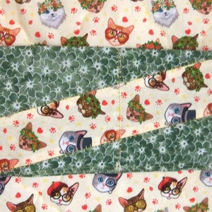 K&ouml;nnte beinhalten: Eine gesteppte Stoffh&uuml;lle mit einem sich wiederholenden Muster aus Katzen, die verschiedene Accessoires wie Blumenkr&auml;nze, Brillen und H&uuml;te tragen, durchsetzt mit kleinen roten Pfotenabdr&uuml;cken. Das Design enth&auml;lt diagonale B&auml;nder aus dunkelgr&uuml;nem Blumenstoff.