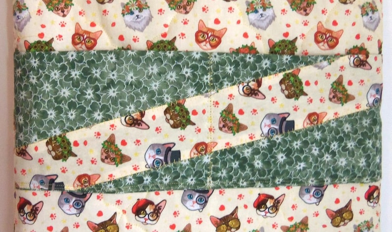 K&ouml;nnte beinhalten: Eine gesteppte Stoffh&uuml;lle mit einem sich wiederholenden Muster aus Katzen, die verschiedene Accessoires wie Blumenkr&auml;nze, Brillen und H&uuml;te tragen, durchsetzt mit kleinen roten Pfotenabdr&uuml;cken. Das Design enth&auml;lt diagonale B&auml;nder aus dunkelgr&uuml;nem Blumenstoff.