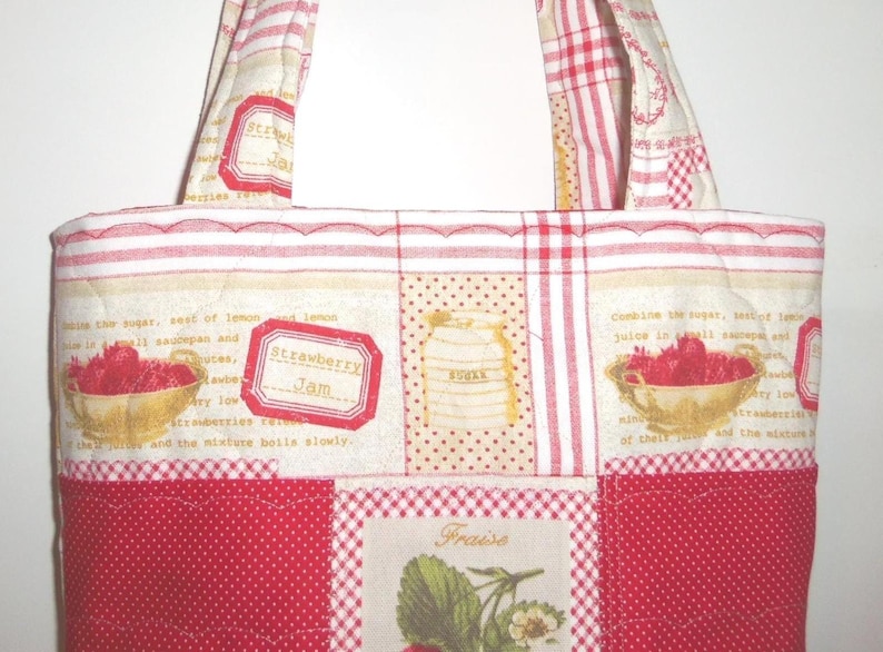 May include: A quilted tote bag featuring a patchwork design with red and white gingham, polka dots, and cream fabric panels. The print includes illustrations of strawberries in bowls, sugar jars, and text reading Strawberry Jam and Fraise.