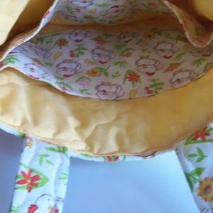 May include: A fabric tote bag with a yellow interior lining and a white exterior featuring a repeating pattern of chickens, green leaves, and red and yellow flowers. The bag includes two fabric handles and an internal pocket with the same print.