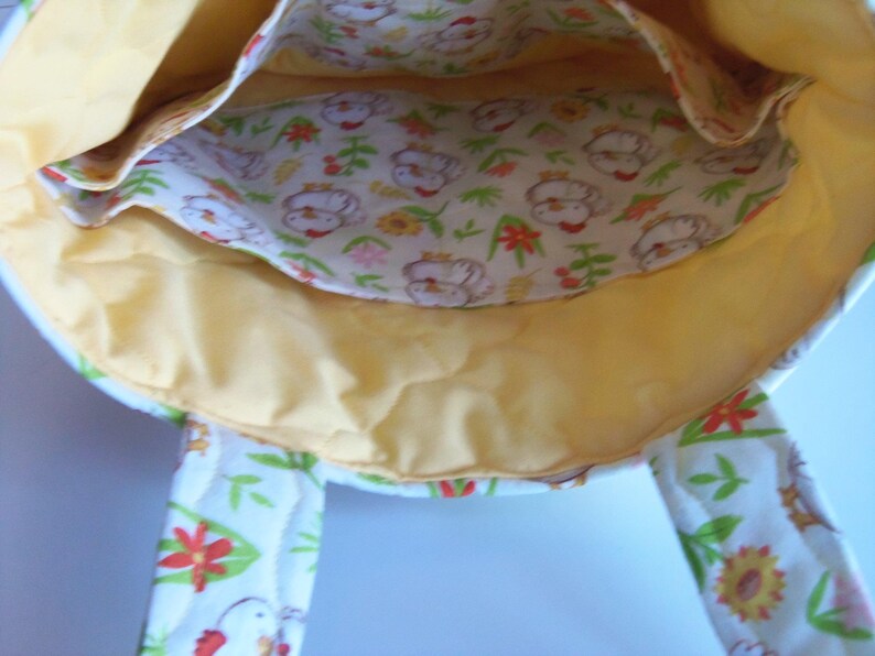 May include: A fabric tote bag with a yellow interior lining and a white exterior featuring a repeating pattern of chickens, green leaves, and red and yellow flowers. The bag includes two fabric handles and an internal pocket with the same print.