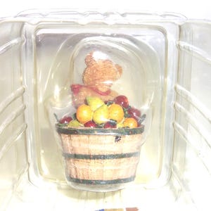 Op de afbeelding: Een beeldje van een teddybeer met een rode sjaal en een witte hoed, zittend achter een houten mand vol fruit. De mand bevat sinaasappels, peren en rode bessen. Het beeldje zit in een doorzichtige plastic verpakking.