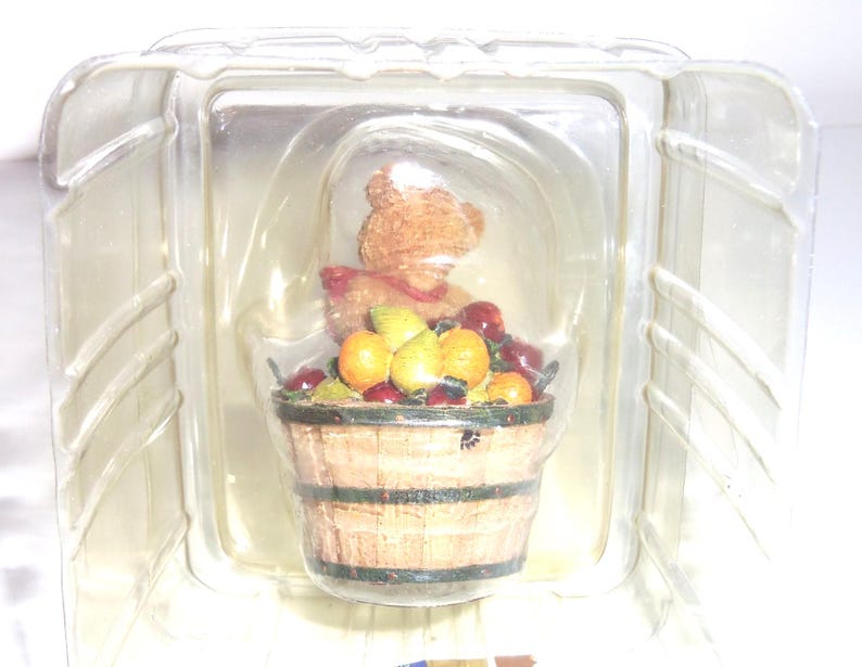 Op de afbeelding: Een beeldje van een teddybeer met een rode sjaal en een witte hoed, zittend achter een houten mand vol fruit. De mand bevat sinaasappels, peren en rode bessen. Het beeldje zit in een doorzichtige plastic verpakking.