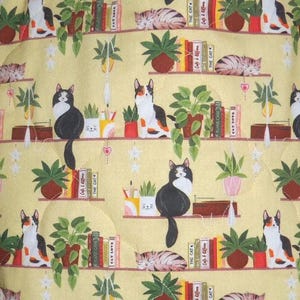 May include: A yellow fabric featuring a repeating pattern of cats, potted plants, and books on shelves. The cats are depicted in various positions, including sitting and sleeping. The books have spines labeled with text such as The Cat and Cat Love.