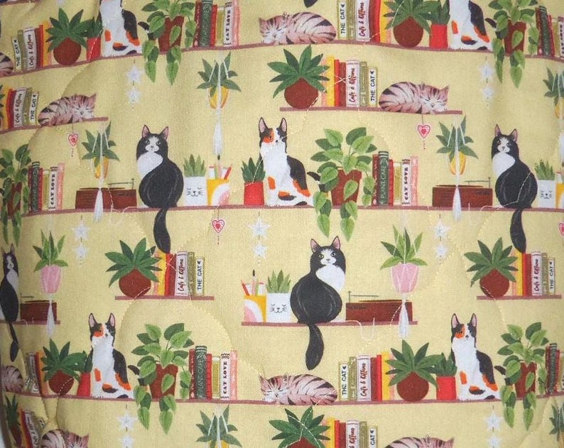May include: A yellow fabric featuring a repeating pattern of cats, potted plants, and books on shelves. The cats are depicted in various positions, including sitting and sleeping. The books have spines labeled with text such as The Cat and Cat Love.