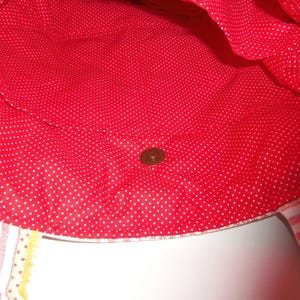 May include: A tote bag interior featuring a bright red fabric lining with a white polka dot pattern. The bag includes a centred metallic magnetic snap closure. Two fabric straps are visible at the bottom, one with a striped pattern.