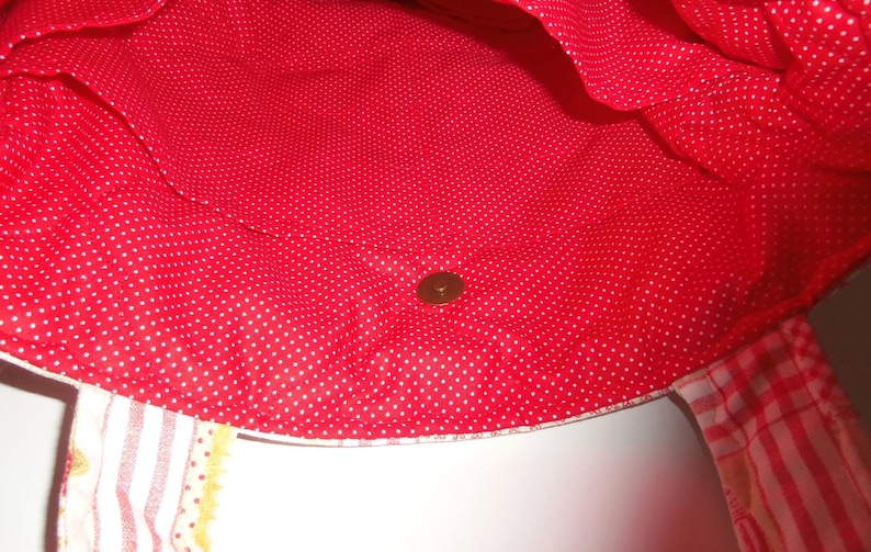 May include: A tote bag interior featuring a bright red fabric lining with a white polka dot pattern. The bag includes a centred metallic magnetic snap closure. Two fabric straps are visible at the bottom, one with a striped pattern.