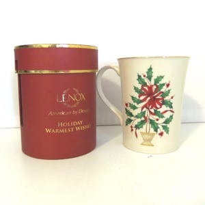 May include: A cream-colored ceramic mug featuring a green holly and red ribbon design, accompanied by a cylindrical red gift box with gold trim and the text LENOX American by Design HOLIDAY WARMEST WISHES printed in gold lettering.