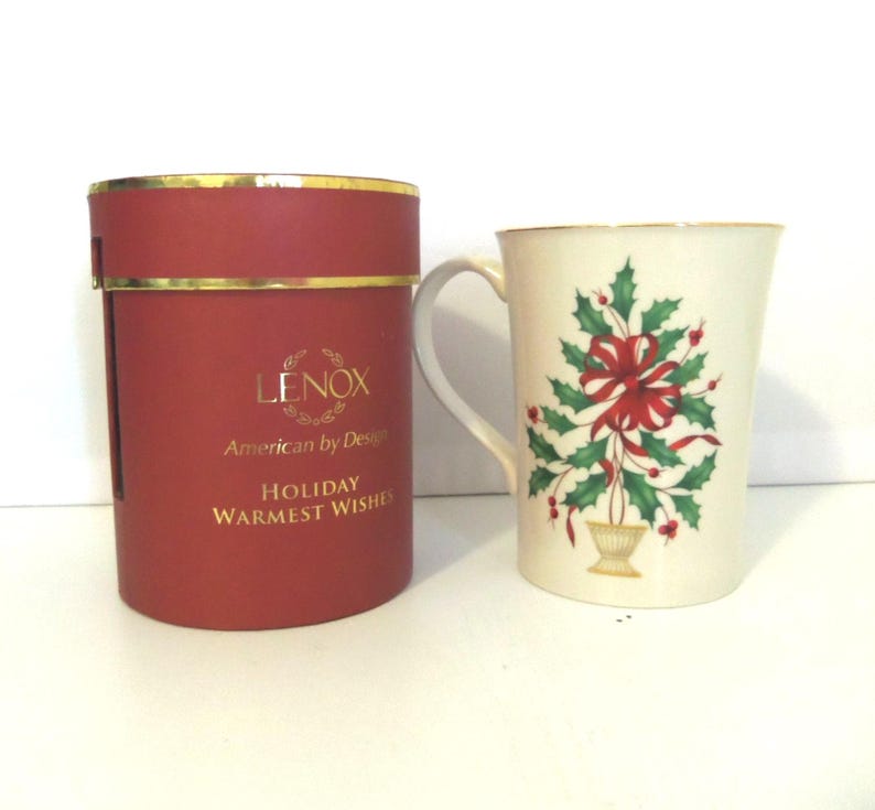 May include: A cream-colored ceramic mug featuring a green holly and red ribbon design, accompanied by a cylindrical red gift box with gold trim and the text LENOX American by Design HOLIDAY WARMEST WISHES printed in gold lettering.