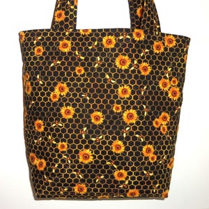 May include: A quilted tote bag featuring a black fabric base with a repeating pattern of yellow sunflowers, orange honeycombs, and small bees. The bag includes two matching fabric handles and a diamond-stitched quilted texture throughout the surface.