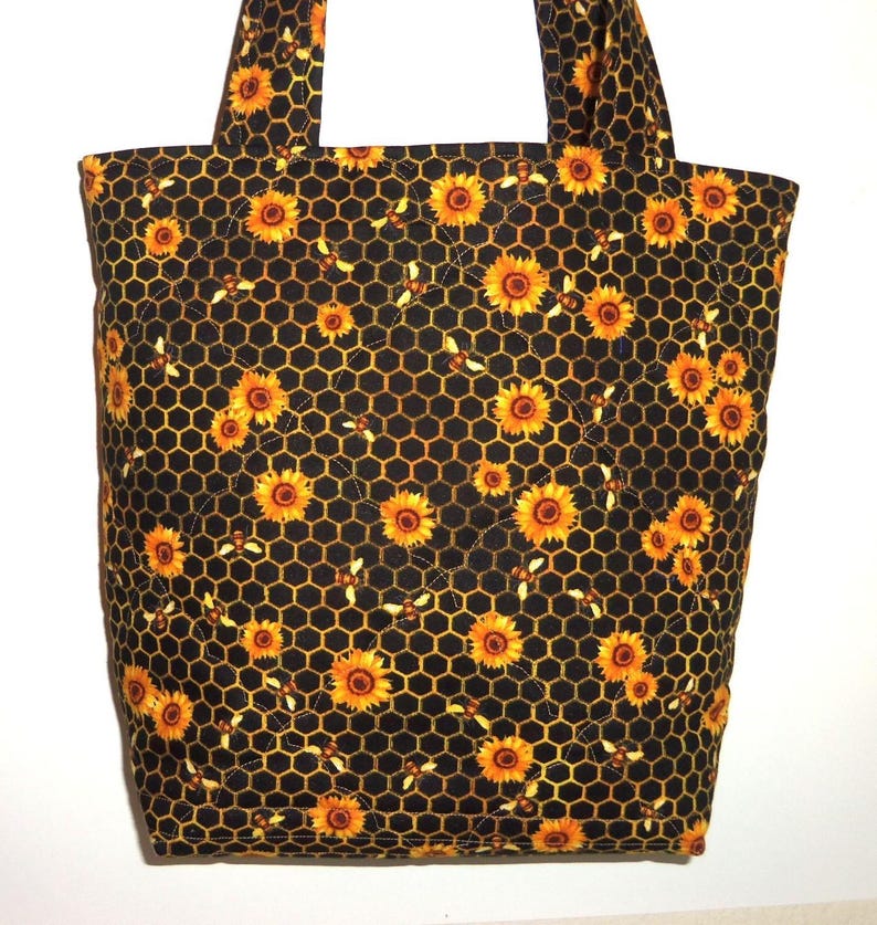 May include: A quilted tote bag featuring a black fabric base with a repeating pattern of yellow sunflowers, orange honeycombs, and small bees. The bag includes two matching fabric handles and a diamond-stitched quilted texture throughout the surface.