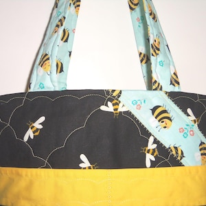 K&ouml;nnte beinhalten: Eine gesteppte Tragetasche mit einem schwarzen Stoffeinsatz, der ein wolkenartiges Steppmuster und Bienenillustrationen aufweist, akzentuiert durch einen hellblauen diagonalen Streifen mit Bienen und Blumen sowie ein gelbes Band am unteren Rand.