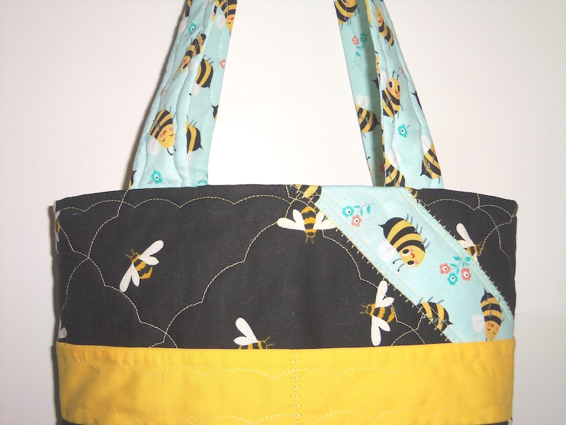 K&ouml;nnte beinhalten: Eine gesteppte Tragetasche mit einem schwarzen Stoffeinsatz, der ein wolkenartiges Steppmuster und Bienenillustrationen aufweist, akzentuiert durch einen hellblauen diagonalen Streifen mit Bienen und Blumen sowie ein gelbes Band am unteren Rand.