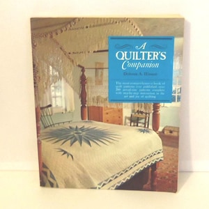 Puede incluir: Un libro de tapa dura titulado A Quilter's Companion de Dolores A. Hinson. La portada presenta una fotograf&iacute;a de una cama con una colcha blanca con un patr&oacute;n de estrella azul, un marco de cama de madera y un cuadro de texto azul con letras blancas.