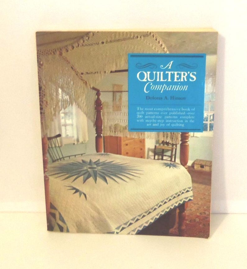Puede incluir: Un libro de tapa dura titulado A Quilter's Companion de Dolores A. Hinson. La portada presenta una fotograf&iacute;a de una cama con una colcha blanca con un patr&oacute;n de estrella azul, un marco de cama de madera y un cuadro de texto azul con letras blancas.