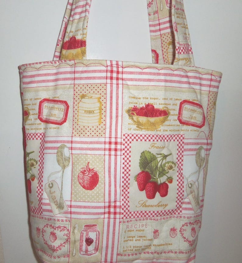 May include: A quilted tote bag featuring a cream-coloured fabric with a red plaid grid pattern. The print includes illustrations of strawberries, jars of jam, sugar canisters, spoons, and text related to strawberry jam recipes and ingredients.