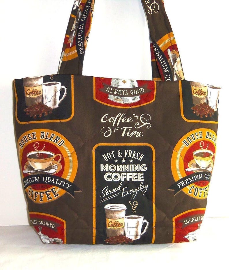 Puede incluir: Un bolso tote acolchado de color marr&oacute;n oscuro con un patr&oacute;n repetitivo de tem&aacute;tica cafetera. El estampado incluye varias tazas de caf&eacute;, granos y textos como "Coffee Time", "House Blend" y "Hot & Fresh Morning Coffee Served Everyday".