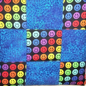 Puede incluir: Un cuadrado de tela acolchada con un patr&oacute;n de cuadr&iacute;cula de bloques alternos. Algunos bloques muestran un fondo negro con un patr&oacute;n repetitivo de caras sonrientes multicolores en rojo, naranja, amarillo, verde, azul y morado. Otros son azules.