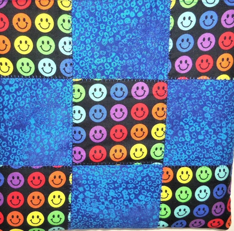 Puede incluir: Un cuadrado de tela acolchada con un patr&oacute;n de cuadr&iacute;cula de bloques alternos. Algunos bloques muestran un fondo negro con un patr&oacute;n repetitivo de caras sonrientes multicolores en rojo, naranja, amarillo, verde, azul y morado. Otros son azules.