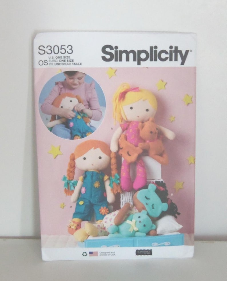 May include: Simplicity sewing pattern S3053 featuring doll designs. The cover shows several handmade dolls with various hairstyles and outfits, including a doll with pigtails and a doll with a yellow bow. The background is pink with yellow star accents.