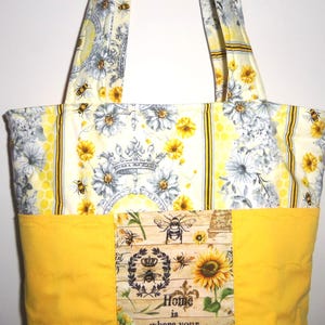 May include: A quilted tote bag with a bee and floral design. The bag features a yellow base and handles, with a patterned upper section showcasing bees, flowers, and a crown. The bag includes the text "Home is where your..."
