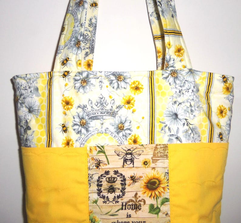 May include: A quilted tote bag with a bee and floral design. The bag features a yellow base and handles, with a patterned upper section showcasing bees, flowers, and a crown. The bag includes the text "Home is where your..."