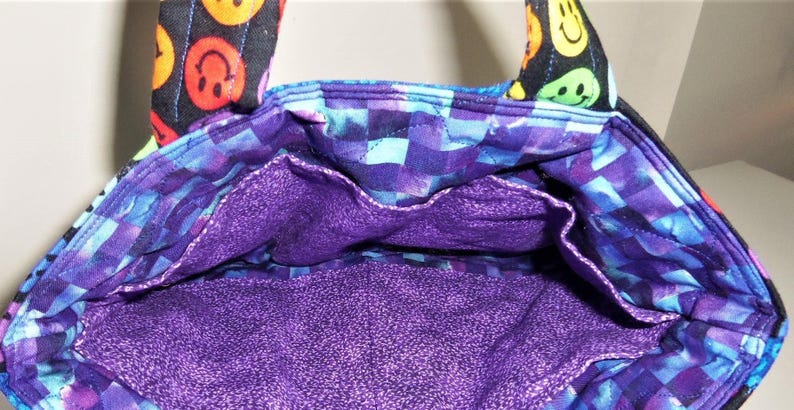 Puede incluir: Un bolso de tela con exterior negro y un patr&oacute;n de caras sonrientes de colores en las asas. El interior est&aacute; forrado con una tela de cuadros morados y azules y una base morada s&oacute;lida, incluyendo un bolsillo abierto en la pared interior.
