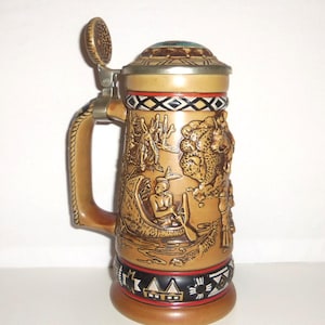 May include: A tan ceramic beer stein featuring a metal lid and a handle. The body displays embossed scenes of a person in a canoe and a buffalo, with decorative black, white, and red geometric patterns around the top and bottom rims.