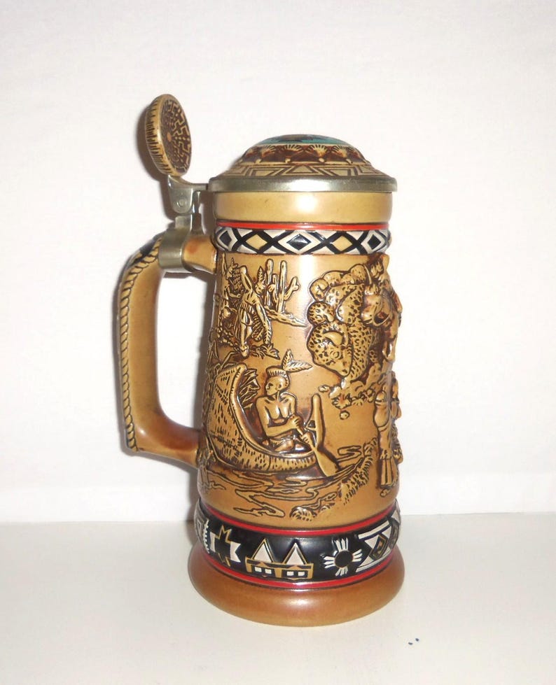 May include: A tan ceramic beer stein featuring a metal lid and a handle. The body displays embossed scenes of a person in a canoe and a buffalo, with decorative black, white, and red geometric patterns around the top and bottom rims.