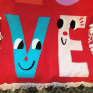 May include: Red fleece blanket with the word "VE" in blue and white, each letter designed with cartoon faces. The blanket has a fringed edge in pink and white.