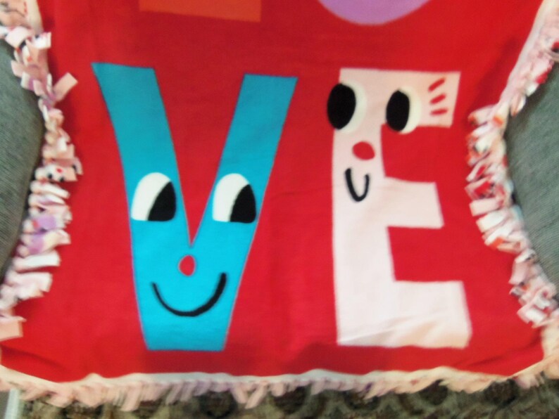 May include: Red fleece blanket with the word "VE" in blue and white, each letter designed with cartoon faces. The blanket has a fringed edge in pink and white.