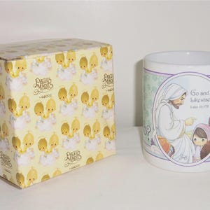 May include: A white ceramic mug featuring a Precious Moments illustration of a figure in robes with children and the text Go and Do likewise. Luke 10:37B. Beside it sits a yellow cardboard box with a repeating pattern of Precious Moments characters.