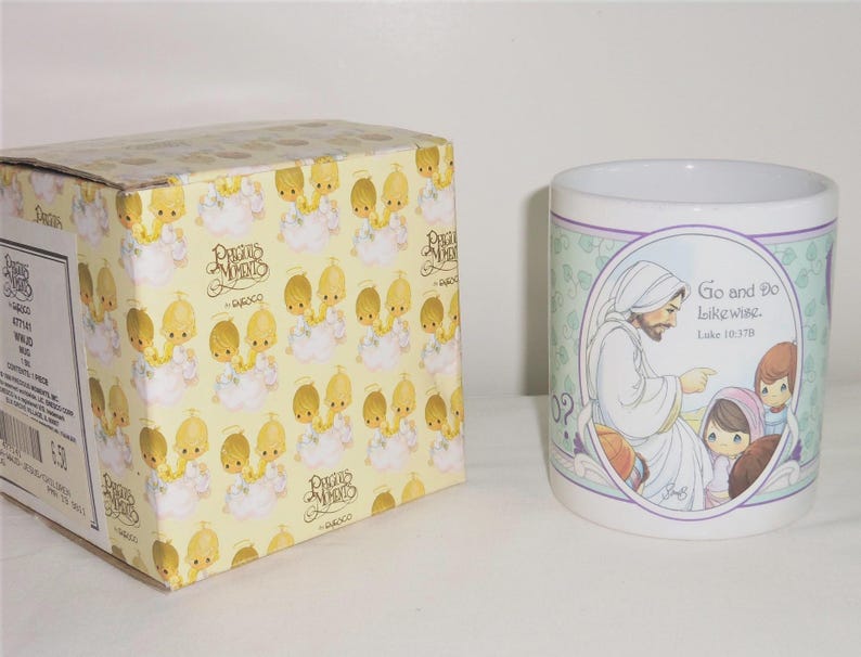 May include: A white ceramic mug featuring a Precious Moments illustration of a figure in robes with children and the text Go and Do likewise. Luke 10:37B. Beside it sits a yellow cardboard box with a repeating pattern of Precious Moments characters.