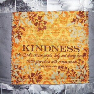 May include: A fabric item with a central panel featuring the word "KINDNESS" in brown text, along with a quote from Colossians 3:12. The panel has an orange floral pattern and is bordered by gray fabric with decorative stitching. The background has a black and white pattern.