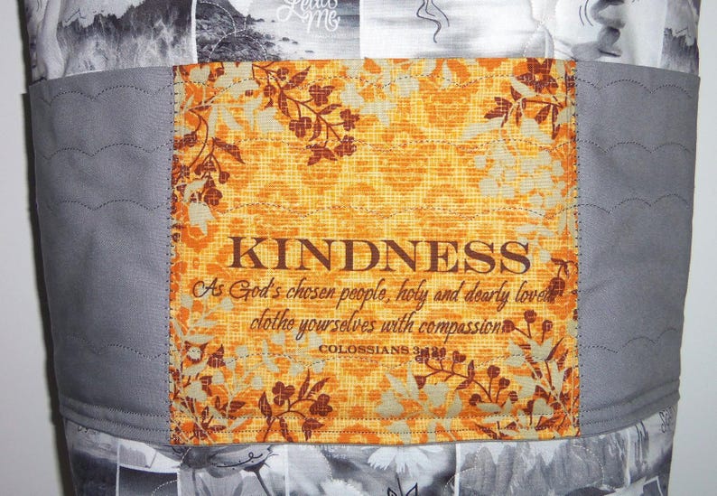May include: A fabric item with a central panel featuring the word "KINDNESS" in brown text, along with a quote from Colossians 3:12. The panel has an orange floral pattern and is bordered by gray fabric with decorative stitching. The background has a black and white pattern.