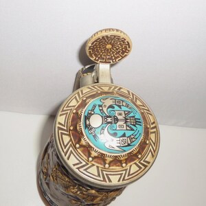 May include: A ceramic beer stein featuring a metal lid with a circular inlay. The inlay displays a turquoise center with a black graphic of two birds facing each other. The lid rim has a cream and brown geometric pattern, and the thumb lift is circular.