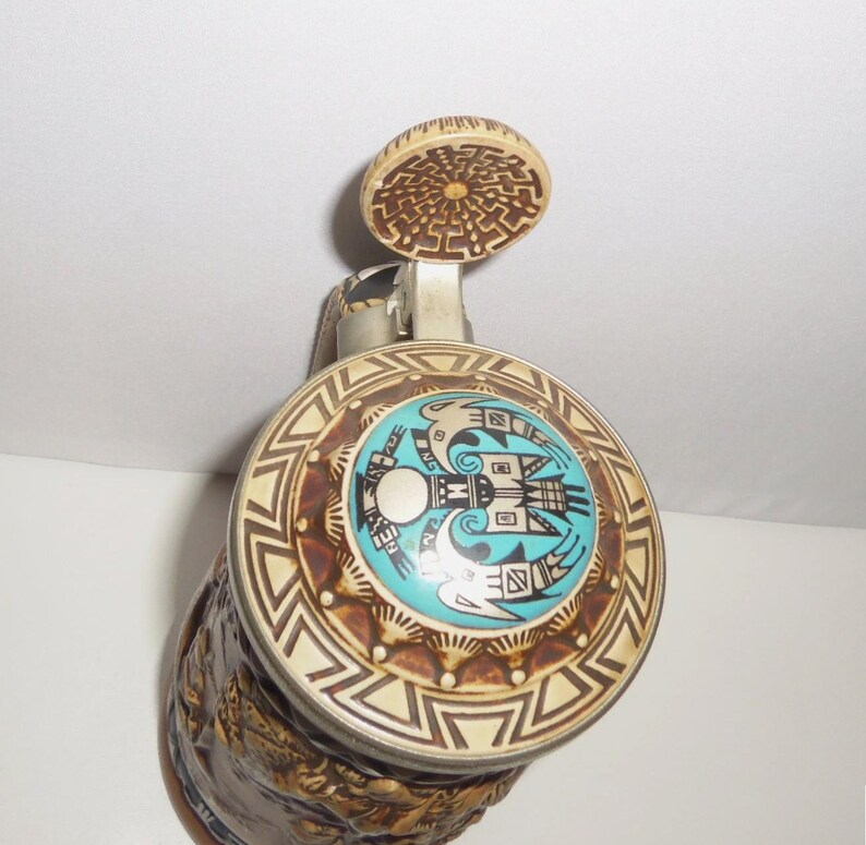 May include: A ceramic beer stein featuring a metal lid with a circular inlay. The inlay displays a turquoise center with a black graphic of two birds facing each other. The lid rim has a cream and brown geometric pattern, and the thumb lift is circular.