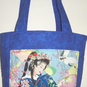 May include: A quilted tote bag featuring a vibrant blue fabric border and handles. The central panel displays a detailed illustration of a person with dark hair adorned with floral accessories, wearing a blue patterned garment amidst cherry blossoms.