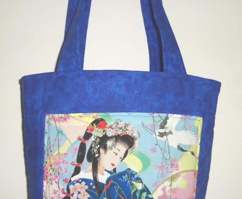 May include: A quilted tote bag featuring a vibrant blue fabric border and handles. The central panel displays a detailed illustration of a person with dark hair adorned with floral accessories, wearing a blue patterned garment amidst cherry blossoms.