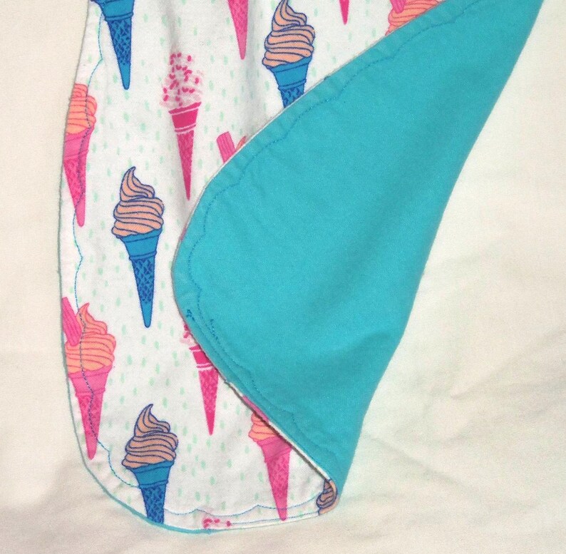 Op de afbeelding: Een babydeken met een patroon van roze, oranje en blauwe ijsjes op een witte achtergrond. De deken heeft een turquoise blauwe achterkant en een geschulpte rand.