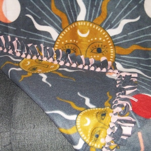 May include: A gray fleece blanket with a repeating pattern of suns and moons. The suns are yellow with a crescent moon inside and have a stylized face with eyes and a nose. The moons are white with pink accents. The blanket has a pink and gray knotted fringe border.